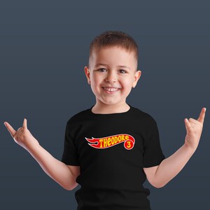 K&ouml;nnte beinhalten: Ein schwarzes T-Shirt mit dem Namen "THEODORE" in einem rot-gelben Design, mit der Zahl "3" in einem Kreis. Das T-Shirt wird von einem l&auml;chelnden Kind getragen.
