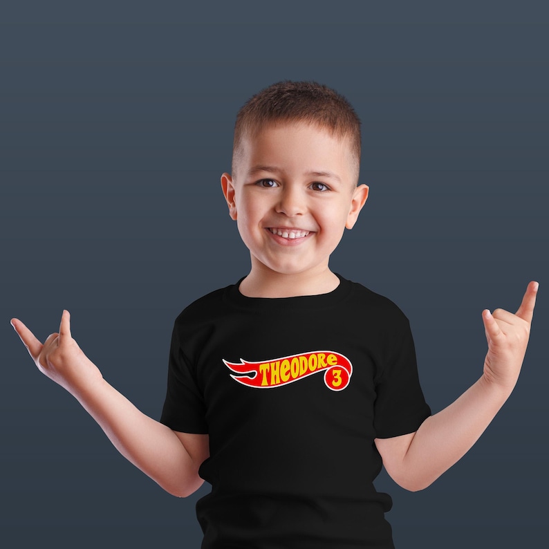 K&ouml;nnte beinhalten: Ein schwarzes T-Shirt mit dem Namen "THEODORE" in einem rot-gelben Design, mit der Zahl "3" in einem Kreis. Das T-Shirt wird von einem l&auml;chelnden Kind getragen.