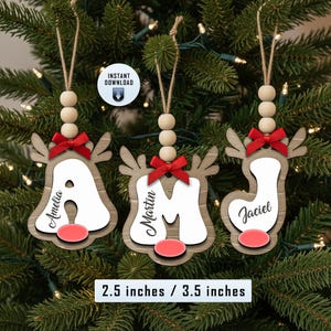 Könnte beinhalten: Drei Weihnachtsornamente aus Holz in Form von Rentieren, jedes mit einem Buchstaben und einem Namen. Sie haben rote Schleifen, rote Nasen und sind mit Schnur und Holzperlen aufgehängt. Die Ornamente sind 6,35 cm x 8,89 cm groß.