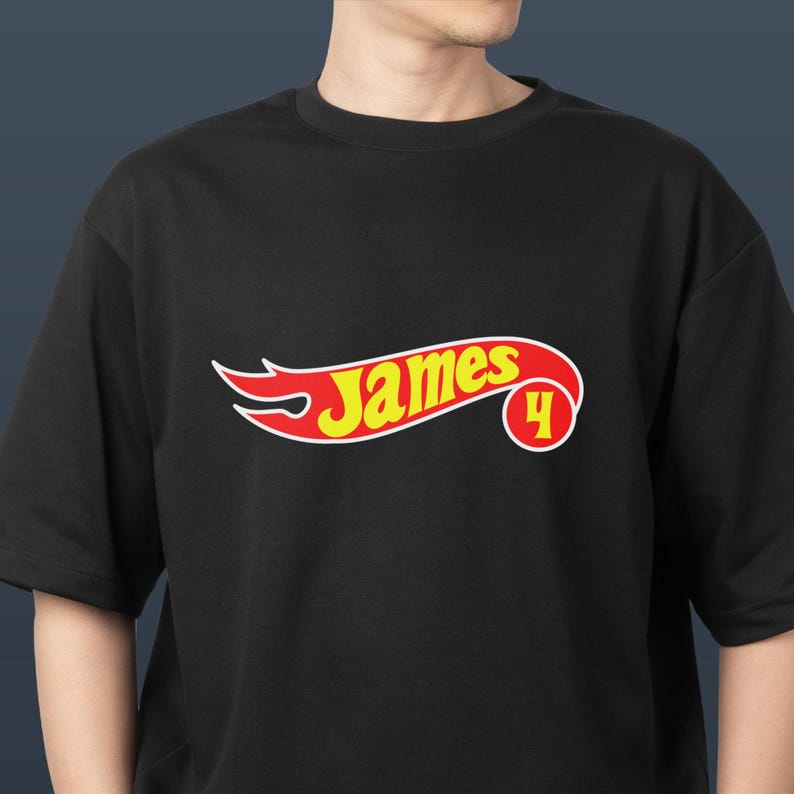 K&ouml;nnte beinhalten: Schwarzes T-Shirt mit einem stilisierten Logo. Der Name "James" ist gelb, wei&szlig; umrandet, mit roten Flammen. Die Zahl "4" befindet sich in einem roten Kreis. Das T-Shirt hat einen Rundhalsausschnitt.