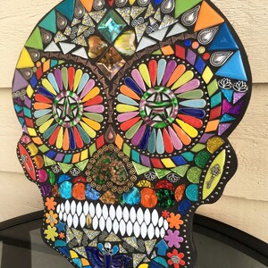 May include: A colorful mosaic skull with a black border. The mosaic features a variety of shapes and colors, including blue, green, orange, yellow, and purple. The skull's teeth are white, and the eyes are decorated with starburst patterns.