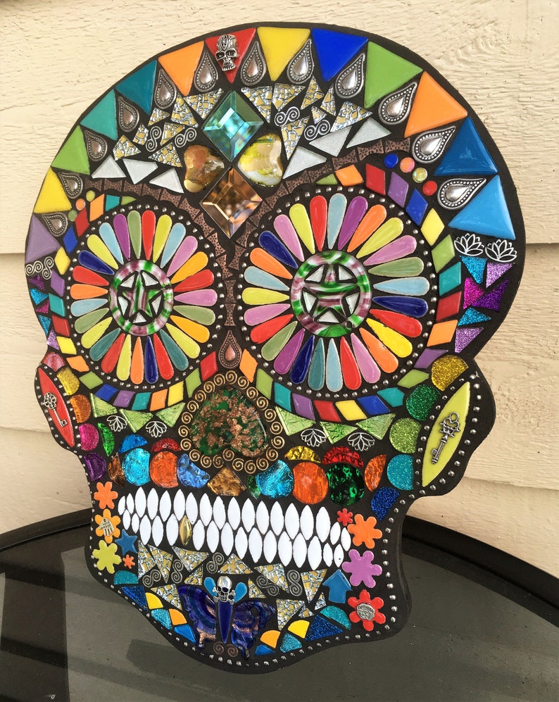 May include: A colorful mosaic skull with a black border. The mosaic features a variety of shapes and colors, including blue, green, orange, yellow, and purple. The skull's teeth are white, and the eyes are decorated with starburst patterns.