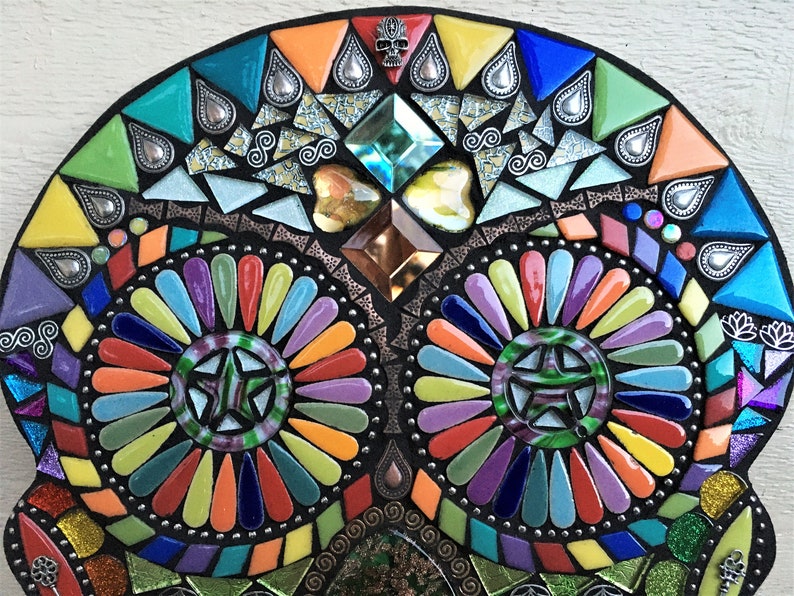 May include: A colorful mosaic skull design with a black background. The mosaic features a variety of shapes and colors, including triangles, diamonds, and teardrops. The design incorporates a star in the eye sockets and a small skull at the top.