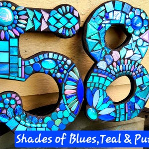 K&ouml;nnte beinhalten: Zwei gro&szlig;e Hausnummern, 5 und 8, verziert mit einem Mosaik aus Glasfliesen in Blau-, T&uuml;rkis- und Violettt&ouml;nen. Die Zahlen weisen verschiedene Fliesenformen auf, darunter runde Edelsteine und geometrische St&uuml;cke, die auf einer dunklen Basis angebracht sind. Der Text lautet: Shades of Blues, Teal & Purple.