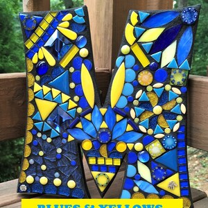 Może przedstawiać: Litera M wykonana z mozaiki z niebieskich i ż&oacute;łtych szklanych płytek. Litera znajduje się na czarnym tle, a pod nią widnieje napis "Blues & Yellows".