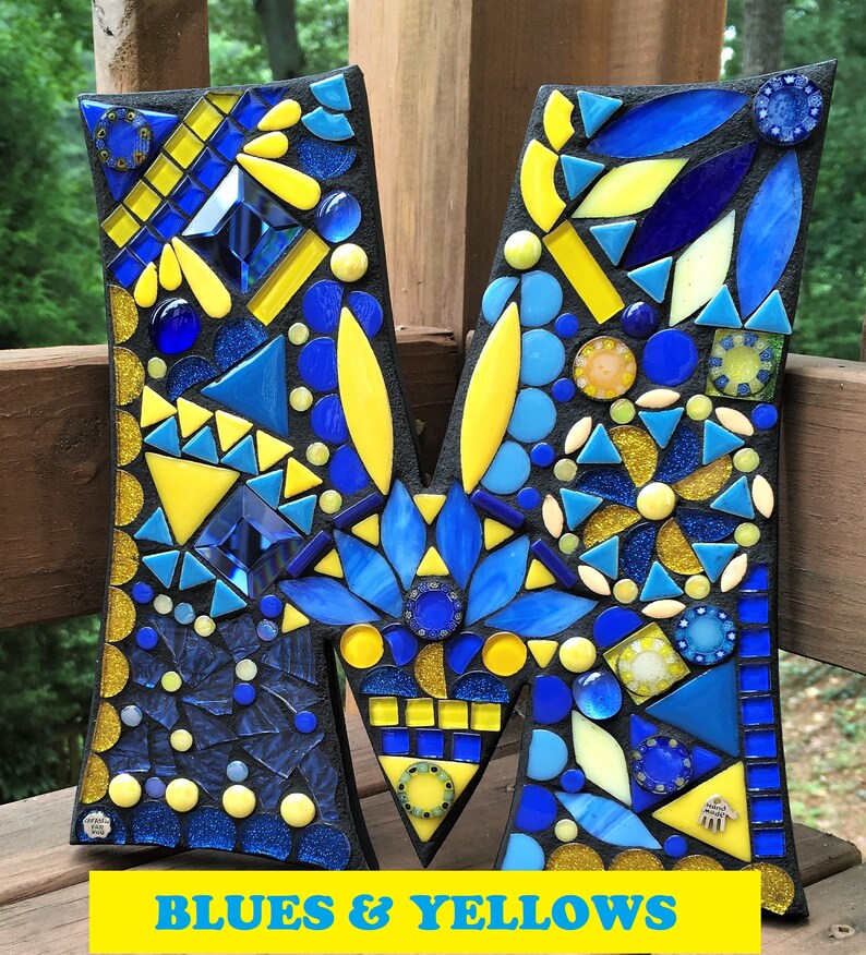 Może przedstawiać: Litera M wykonana z mozaiki z niebieskich i ż&oacute;łtych szklanych płytek. Litera znajduje się na czarnym tle, a pod nią widnieje napis "Blues & Yellows".