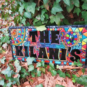 May include: A rectangular mosaic sign with the text "THE KILANIS" in black letters. The sign features colorful mosaic patterns in red, blue, yellow, and green. The sign is surrounded by green ivy.
