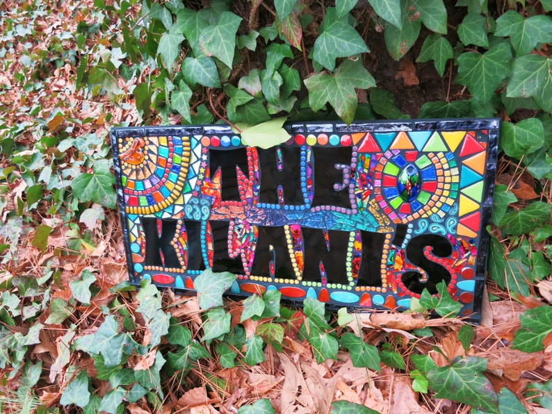 May include: A rectangular mosaic sign with the text "THE KILANIS" in black letters. The sign features colorful mosaic patterns in red, blue, yellow, and green. The sign is surrounded by green ivy.