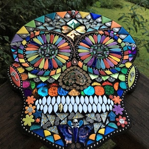 May include: A colorful mosaic skull with intricate details. The mosaic features a variety of shapes and colors, including orange, blue, green, and yellow. The skull has large eye sockets and a row of white teeth. Decorative keys and flowers are also included.