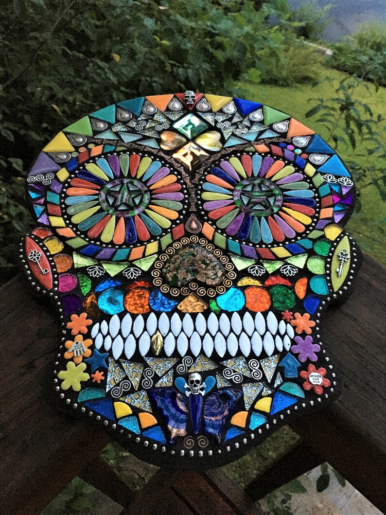 May include: A colorful mosaic skull with intricate details. The mosaic features a variety of shapes and colors, including orange, blue, green, and yellow. The skull has large eye sockets and a row of white teeth. Decorative keys and flowers are also included.