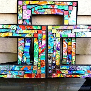 May include: A set of three rectangular picture frames arranged in a cluster, each featuring a mosaic surface composed of small, multi-colored glass tiles in shades of blue, green, yellow, orange, pink, and red, set within a black frame border.