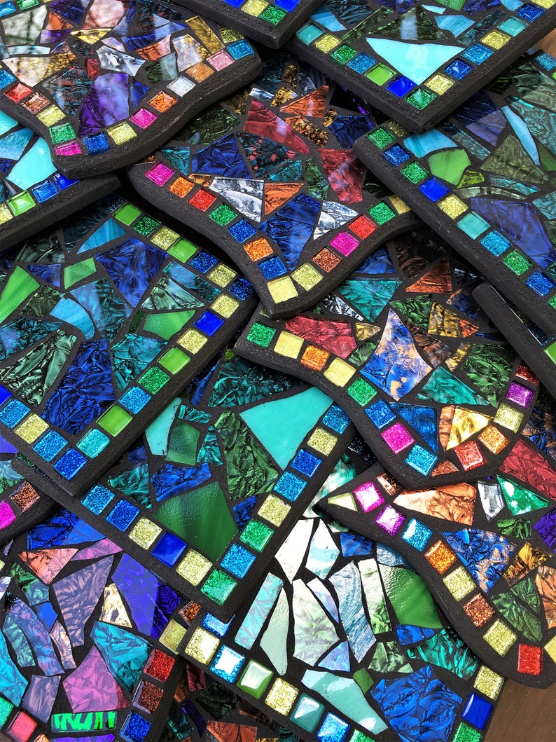 K&ouml;nnte beinhalten: Ein Stapel bunter Mosaik-Untersetzer mit schwarzem Rand. Die Untersetzer bestehen aus kleinen Glassplittern in Blaut&ouml;nen, Gr&uuml;n, Rot und Gelb.