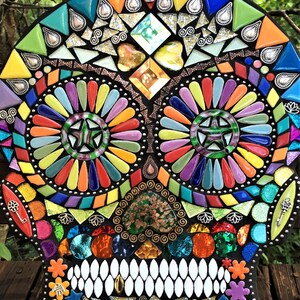May include: A colorful mosaic skull with a black background. The skull features vibrant hues of yellow, orange, red, green, blue, and purple. The eyes are circular with a star design. The teeth are white, and the design includes decorative elements.