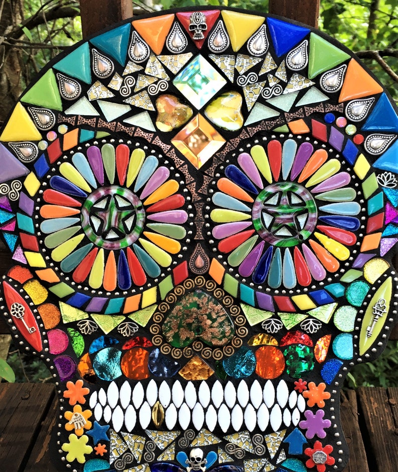 May include: A colorful mosaic skull with a black background. The skull features vibrant hues of yellow, orange, red, green, blue, and purple. The eyes are circular with a star design. The teeth are white, and the design includes decorative elements.