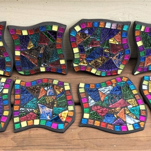 K&ouml;nnte beinhalten: Acht bunte Mosaik-Untersetzer mit schwarzem Rand und wellenf&ouml;rmigem Rand. Jeder Untersetzer zeigt ein einzigartiges Muster aus irisierenden Glasfliesen in Blaut&ouml;nen, Gr&uuml;n, Rot, Gelb und Lila.