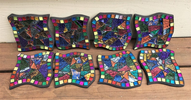 K&ouml;nnte beinhalten: Acht bunte Mosaik-Untersetzer mit schwarzem Rand und wellenf&ouml;rmigem Rand. Jeder Untersetzer zeigt ein einzigartiges Muster aus irisierenden Glasfliesen in Blaut&ouml;nen, Gr&uuml;n, Rot, Gelb und Lila.