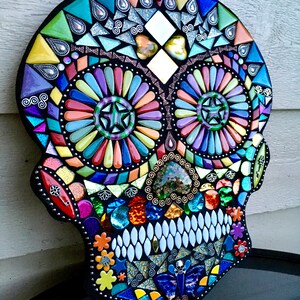 May include: A colorful mosaic skull with a black border. The mosaic features a variety of shapes and colors, including blue, green, orange, and purple. The eyes are circular with star-shaped centers. The teeth are white, and the overall design is intricate.