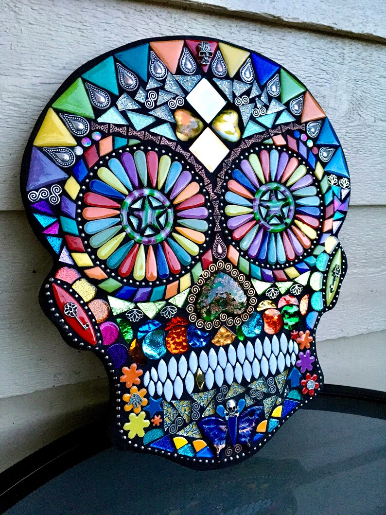 May include: A colorful mosaic skull with a black border. The mosaic features a variety of shapes and colors, including blue, green, orange, and purple. The eyes are circular with star-shaped centers. The teeth are white, and the overall design is intricate.