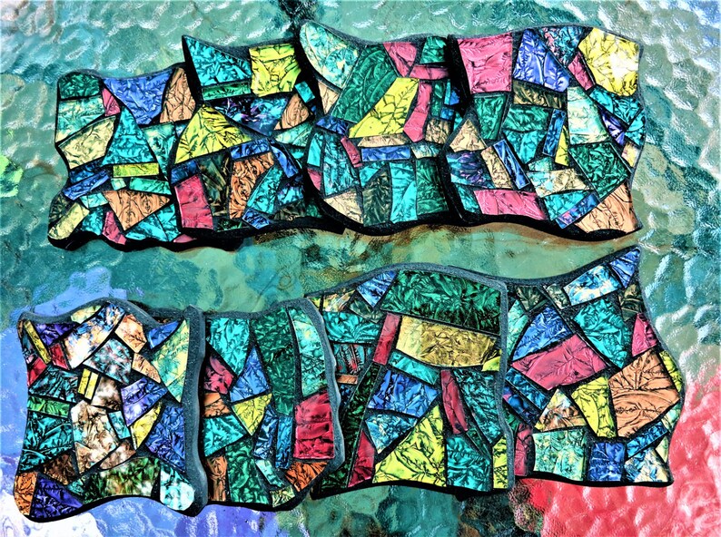K&ouml;nnte beinhalten: Ein Set aus acht bunten Mosaik-Untersetzern mit geometrischem Design. Die Untersetzer sind aus Glas und haben einen schwarzen Rand. Sie sind in zwei Reihen zu je vier angeordnet.
