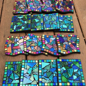 K&ouml;nnte beinhalten: Ein Set mit 12 bunten Mosaik-Untersetzern mit verschiedenen geometrischen Mustern. Die Untersetzer bestehen aus kleinen Glassplittern und haben einen schwarzen Rand.
