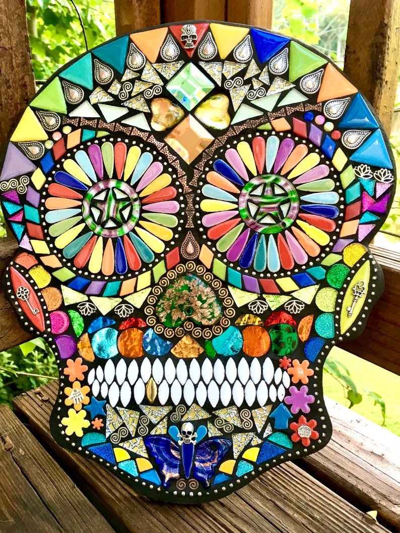 May include: A colorful mosaic skull with a black border. The mosaic features a variety of shapes and colors, including blue, green, yellow, orange, and purple. The eyes are circular with star-shaped centers. Decorative keys and a butterfly are also included.