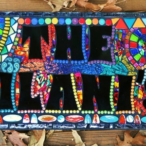 May include: A rectangular mosaic sign with the words "THE KILANI'S" in large black letters. The sign is framed with a dark blue border and features colorful mosaic tiles in various shapes and colors, including red, yellow, blue, and green.