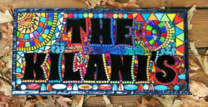 May include: A rectangular mosaic sign with the words "THE KILANI'S" in large black letters. The sign is framed with a dark blue border and features colorful mosaic tiles in various shapes and colors, including red, yellow, blue, and green.