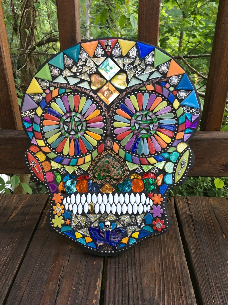 May include: A colorful mosaic skull with intricate details. The artwork features a variety of glass tiles in shades of orange, yellow, green, blue, and purple. The skull's eyes are designed with star-shaped patterns. The mosaic is displayed on a wooden surface.