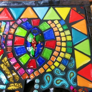 May include: A colorful mosaic art piece with a black background. The mosaic features a variety of shapes, including triangles, squares, and oval shapes, in vibrant colors like red, orange, yellow, green, and blue. The design includes paisley patterns and glitter accents.