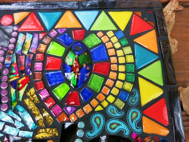 May include: A colorful mosaic art piece with a black background. The mosaic features a variety of shapes, including triangles, squares, and oval shapes, in vibrant colors like red, orange, yellow, green, and blue. The design includes paisley patterns and glitter accents.