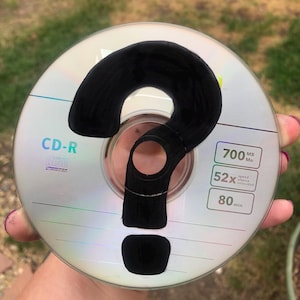 Puede incluir: Un disco CD-R con un gran signo de interrogación negro dibujado. El disco tiene el texto "CD-R" y especificaciones de 700 MB, velocidad 52x y 80 minutos. El disco está sostenido en una mano.
