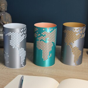 May include: Four cylindrical candle holders with a world map design. The holders are white, silver, teal, and silver with blue, white, peach, and gold interiors. The map is created with a honeycomb pattern. A notebook and pen are in the foreground.