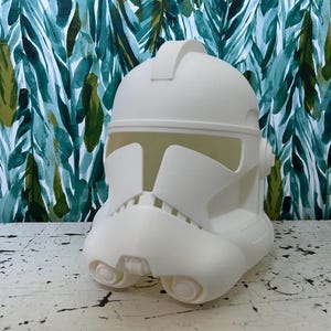 Puede incluir: Un casco blanco de soldado clon de Star Wars de plástico. El casco presenta un diseño distintivo con una visera, un aparato respiratorio y otros detalles. El casco está sobre una superficie blanca con un fondo estampado en verde y azul.
