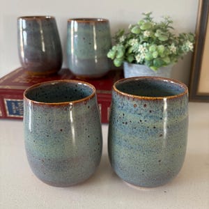 May include: Four handmade ceramic tumblers with a speckled blue-green glaze and a brown rim. The tumblers have a smooth, rounded shape. A small potted plant is visible in the background. The tumblers are arranged on a white surface.