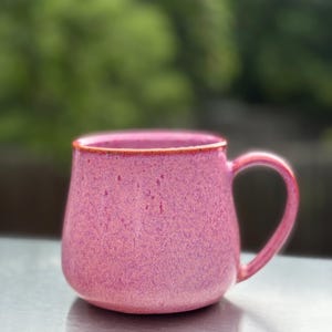 May include: A rounded ceramic mug with a speckled pink finish and a smooth, curved handle. The rim of the mug features a thin, contrasting orange-brown edge, and the overall surface texture appears slightly mottled with darker pink tones.