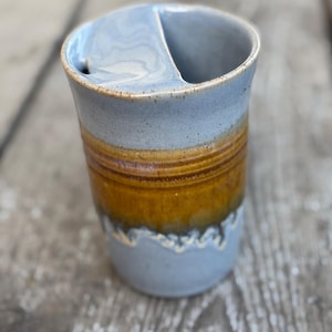 May include: A ceramic tumbler featuring a two-tone glaze with a light blue top and base separated by a wide band of deep amber brown. The cup includes an integrated ceramic lid with a semi-circular opening for drinking, showcasing a smooth finish.