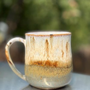 May include: A ceramic mug featuring a reactive glaze with a cream-coloured upper section that transitions into a warm brown drip pattern, followed by a muted olive green base. The mug has a curved handle and a smooth, rounded cylindrical body shape.