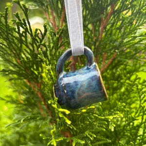 May include: A miniature ceramic mug ornament featuring a glossy, mottled blue and dark navy glaze. The small mug has a curved handle and is suspended by a thin, light gray ribbon, allowing it to hang from a branch of a green coniferous tree.