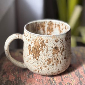 Peut inclure: Une tasse en céramique avec un corps arrondi et une anse incurvée, présentant une couleur de base blanc cassé mouchetée avec des taches brunes texturées et irrégulières réparties sur les surfaces extérieures et intérieures.