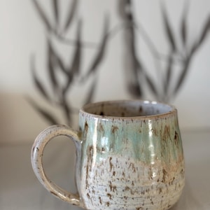 Puede incluir: Una taza de cerámica hecha a mano con un cuerpo redondeado y un asa grande. La taza presenta una base moteada de color blanquecino con un degradado de esmalte verde claro y marrón. La taza mide aproximadamente 10 cm de alto.