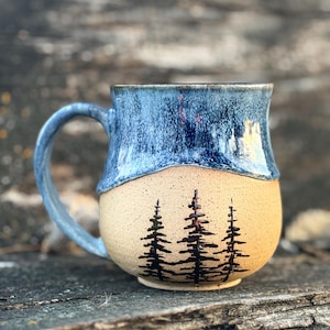 Puede incluir: Una taza de cerámica con diseño bicolor, que presenta un esmalte azul brillante y moteado en la parte superior y una base de color beige claro con motas. La sección inferior está decorada con una ilustración negra pintada a mano de tres pinos.