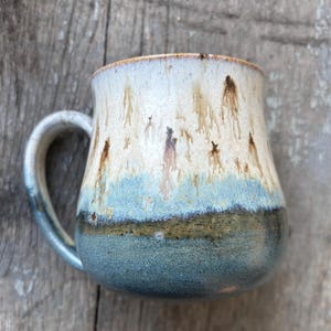 May include: A ceramic mug with a rounded body and a curved handle. The glaze features a gradient transition from a creamy off-white top with brown vertical drip patterns to a mid-section of light blue, ending in a dark, textured forest green base.