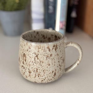 May include: A ceramic mug with a rounded shape and a curved handle. The exterior features a textured, speckled finish with a cream-coloured base and dark brown, irregular spots that create a mottled appearance across the entire surface.