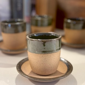 May include: A set of ceramic cups and saucers featuring a two-tone design. The lower portion of each cup and the center of each saucer have a speckled tan finish, while the upper rim and outer edge of the saucers are glazed in dark green.