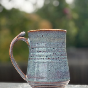 Peut inclure: Une tasse en céramique avec un émail bleu et vert tacheté, présentant des stries horizontales sur la base et le corps. La tasse possède une anse incurvée avec un petit repose-pouce sur le dessus. La finition montre des tons bruns et rouille.