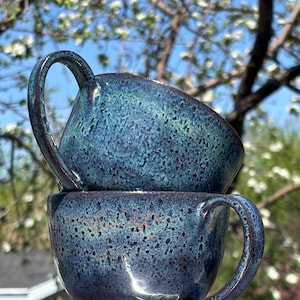 May include: Two handmade ceramic mugs, one stacked on the other. They are a deep blue with a speckled, textured glaze. Each mug has a curved handle. The background shows a blurred tree with white blossoms.