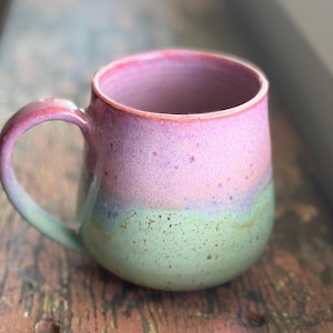 Peut inclure: Une tasse en céramique avec un corps arrondi et une anse incurvée. La partie supérieure de la tasse présente un émail rose vif, tandis que la section inférieure est d'un vert sauge doux avec des taches brun foncé sur toute la surface.