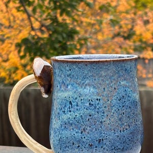 Peut inclure: Une tasse en céramique avec un vernis bleu tacheté et une anse incurvée de couleur claire. Un petit accent brillant en forme de champignon brun foncé est fixé au sommet de l'anse. La tasse présente un corps arrondi et un bord lisse.
