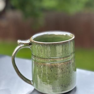 May include: A handmade ceramic mug with a glossy light green glaze featuring subtle brown speckles and horizontal ridges along the lower body. The mug has a sturdy, curved handle attached to the side and a smooth, rounded rim at the top.