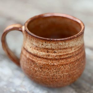 May include: A round ceramic mug with a speckled, textured finish in shades of warm brown and cream. The mug features a wide, bulbous body, a curved handle on the left side, and a smooth, glossy rim that transitions into a darker, earthy tone.